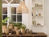 Et hyggeligt rum med potteplanter på et rustikt træbord, flettede kurve, en rattan-pendel og en hvid hylde med vaser, bøger og små dekorationer nær et vindue.