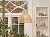 En flettet pendellampe hænger foran et vindue med hvid ramme, og der står grønne planter og dekorative vaser på hylder ved siden af.