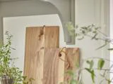 To skærebrætter af træ med læderstropper læner sig op ad en hvid væg, omgivet af grønne planter i et lyst køkken.
