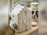 En hvid keramisk husformet lanterne med korsformede vinduer står i vindueskarmen, pyntet med grønne planter og et tændt lys ved siden af.