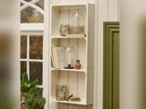 En hvid væghylde med glaskrukker, en lille lerkrukke, en stribet bog og bordskånere ved siden af et vindue og en grøn dør. En potteplante står nedenunder.