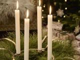 Fire tændte hvide stearinlys står i en krans af grønt løv med bånd, med et pyntet juletræ i baggrunden.