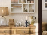 En rustik trækommode med dekorative genstande, herunder en lampe, små trækasser, keramikkande, stearinlys, figurer af hvide heste og en hvid hylde med keramik og ornamenter på en mønstret væg.