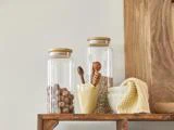 To glaskrukker med trælåg indeholder nødder og frø. Foran står to keramikskåle, træredskaber og et strikket gult klæde, alt sammen på en træhylde mod en lys væg.
