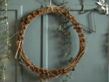 En rustik krans af kviste og små kogler hænger på en væg med blågrønne paneler, og der er grønne planter og lyskæder i nærheden.