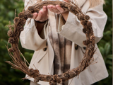En person i beige frakke holder en cirkulær krans af kviste og små pinjekerner, og i baggrunden ses sløret grønt.