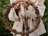 En person i beige frakke holder en cirkulær krans af kviste og små pinjekerner, og i baggrunden ses sløret grønt.