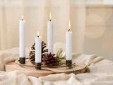 Fire tændte hvide stearinlys står oprejst på en træbakke med kogler og fyrrenåle oven på et draperet beige klæde.