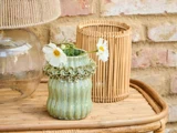 En lille grøn keramikvase med flæsedetaljer indeholder hvide blomster på et kurvebord ved siden af en glaslampe og en bambuslygte med en murstensvæg i baggrunden.