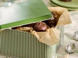 En grønstribet blikæske beklædt med pergamentpapir, delvist tildækket, fyldt med chokoladeovertrukne godbidder med nødder på toppen. Skeer og en tallerken står i nærheden på en ternet dug.