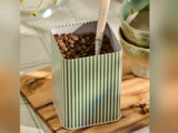 En stribet grøn og hvid dåse fyldt med kaffebønner og en øse med træhåndtag indeni står på en træplade ved siden af stablede keramikskåle.