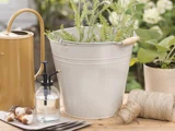 En hvid metalspand med grønne planter, en gul vandkande, en sprayflaske i glas, blomsterløg, sejlgarn og en potteplante på et træbord udendørs.