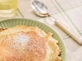 Et nærbillede af en crepe drysset med flormelis på en grøn tallerken med en ske og et glas i baggrunden på en ternet dug.