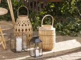 Tre lanterner med lys - to lavet af flettet bambus og en af sort metal og glas - står på en flettet måtte udendørs, omgivet af grønne planter.