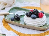 En lille pavlova med brombær, blåbær og hindbær ligger på en grøn tallerken ved siden af en ske på en hæklet dækkeserviet.
