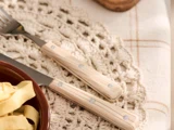 Et nærbillede af en gaffel og en kniv med lyse træskafter, placeret på en hæklet dækkeserviet ved siden af en skål pasta.