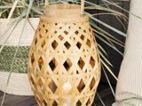 En vævet bambuslygte med håndtag står på en træplade med et tændt hvidt lys indeni. I baggrunden ses dekorativt græs og en pude.
