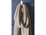 Et tykt, lysegråt strikket tørklæde med struktureret mønster hænger på en hvid knage mod en mørkeblå væg.