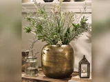 Stor messingvase med forskellige grønne og hvide vilde blomster, ved siden af en lille glaskrukke med vand og en metallanterne med et tændt stearinlys, arrangeret på en træplade.