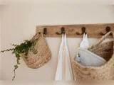 Et vægstativ i træ med metalkroge holder flettede kurve, en plante og hvide håndklæder op mod en beige væg.