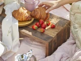 En hånd rækker ud efter en croissant på en rustik træbakke med croissanter, friske jordbær og et lyserødt krus, der står på et let tæppe udendørs.