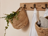 Et vægstativ af træ med tre kroge holder en flettet kurv med grønne blade, et hvidt håndklæde og en anden flettet kurv med gråt stof.