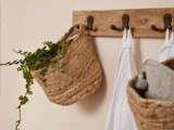 Et vægstativ af træ med tre kroge holder en flettet kurv med grønne blade, et hvidt håndklæde og en anden flettet kurv med gråt stof.