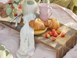 To croissanter på en gul tallerken, friske jordbær, to lyserøde kopper, en hvid serviet og en vase med grønt på en rustik træbakke, der ligger på et lyserødt picnictæppe.