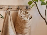 Et beige håndklæde og en flettet kurv med sammenrullede håndklæder hænger på vægkroge ved siden af en grøn plante.