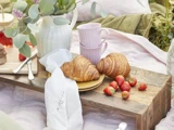 To croissanter og friske jordbær på en gul tallerken ligger på en rustik træbakke med to lyserøde krus og en hvid kande i nærheden, klar til en hyggelig udendørs picnic.