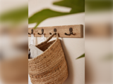 En vævet stråtaske og hvide klude hænger på et vægstativ af træ med metalkroge; grønne blade er slørede i forgrunden.