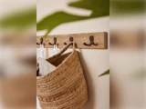 En vævet stråtaske og hvide klude hænger på et vægstativ af træ med metalkroge; grønne blade er slørede i forgrunden.
