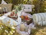 Et hyggeligt picnic-setup på græs med puder, et tæppe, croissanter, jordbær, drikkevarer i flasker, blomster i en vase og en flettet taske omgivet af vilde blomster.