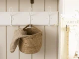 En flettet kurv med et struktureret beige stof indeni hænger på en hvid knagerække, der er monteret på en lys trævæg.