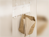 En flettet kurv og et beige tekstureret håndklæde hænger på hvide kroge på en cremefarvet væg.