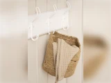 En flettet kurv og et beige tekstureret håndklæde hænger på hvide kroge på en cremefarvet væg.