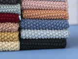 En stak foldede, tykke strikkede stoffer i forskellige farver, bl.a. rød, pink, blå, sort, brun, gul og grå, arrangeret pænt på en blå overflade.