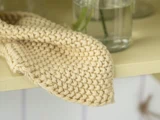 Et nærbillede af et beige hæklet klæde, der er draperet over kanten af en lysegul flade, med glasvaser med vand og grønne stængler i baggrunden.