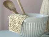 En hvid keramisk røreskål med en beige hæklet karklud draperet over kanten og to træskeer indeni.
