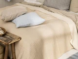 En pænt redt seng med beige og cremefarvede tæpper, en lyseblå pude og puder i neutrale farver ved siden af et rustikt sidebord i træ.