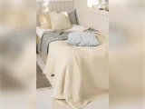 En hyggelig, pænt redt seng med cremefarvede, grå og beige puder og tæpper i et solbeskinnet soveværelse i neutrale farver.