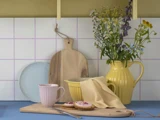 En køkkenscene med et skærebræt, kiks, et lyserødt krus, en gul kande med vilde blomster, en blå tallerken og en gul skål på et blåt bord mod en flisebelagt væg.