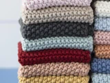 En pænt stablet bunke af strikkede stoffirkanter i forskellige farver, bl.a. pink, grå, blå, rød, beige og gul.