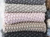 Et nærbillede af fem foldede, hæklede tæpper stablet i neutrale farver: mørkegrå, lysegrå, lyserød, beige og taupe.