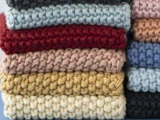 En stak foldede, tyktstrikkede stofstykker i forskellige farver, bl.a. blå, rød, pink, beige, gul, creme og sort.