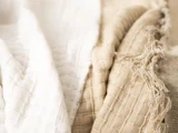 Blødt, struktureret stof i hvide og beige nuancer med synlige vævede mønstre og nogle flossede kanter, arrangeret i blide folder.