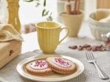 To småkager med pink glasur og krymmel på en tallerken, ved siden af et gult krus og en gaffel, på et hyggeligt dækket bord med varm, blød belysning.