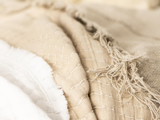 Nærbillede af lagdelte beige og hvide teksturerede stoffer med synlige syninger og flossede kanter, der skaber et blødt og hyggeligt udseende.