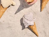 Tre vaffelisvafler på sand, der hver holder en kugleformet kugle af strikket garn i stedet for rigtig is; to hænder er delvist synlige, også iført strikket betræk.