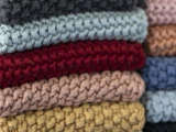 Et nærbillede af stablede, strikkede stoffer i forskellige farver, bl.a. grå, blå, rød, pink og gul. Den strikkede tekstur er tydeligt synlig.
